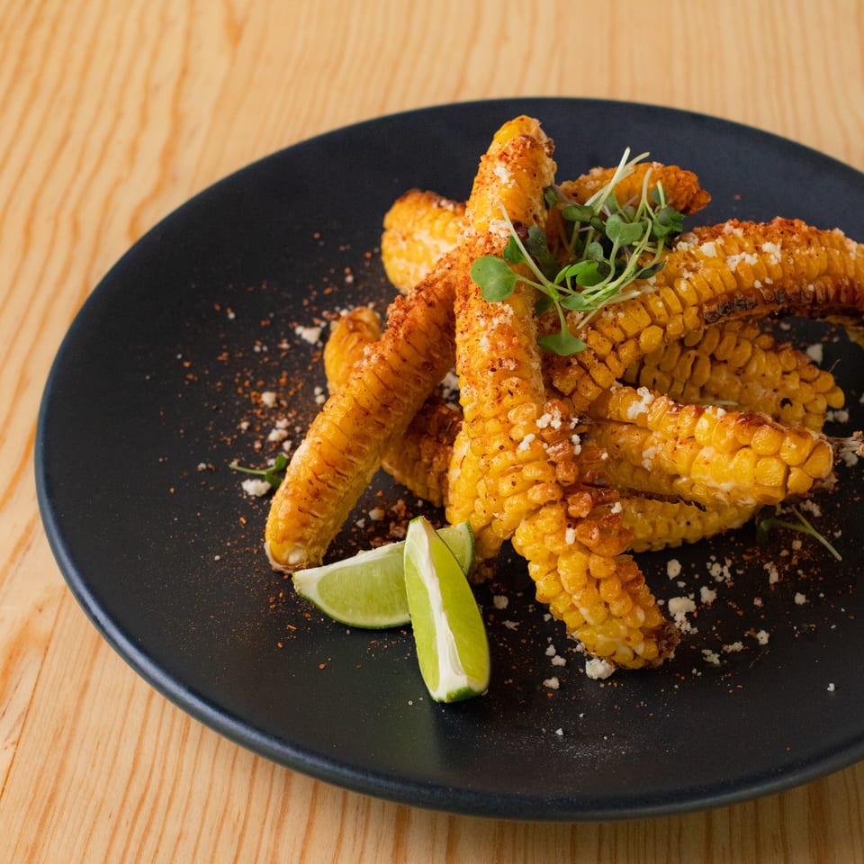 Seasoned grilled corn ribs piled on a black plate with microgreens, lime wedges, crumbled cheese, and spices.