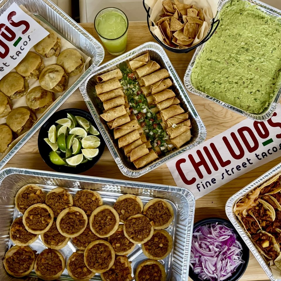 Overhead spread of Chiludos street tacos featuring tacos, taquitos, guacamole, tortilla chips, limes, and salsas in foil trays.
