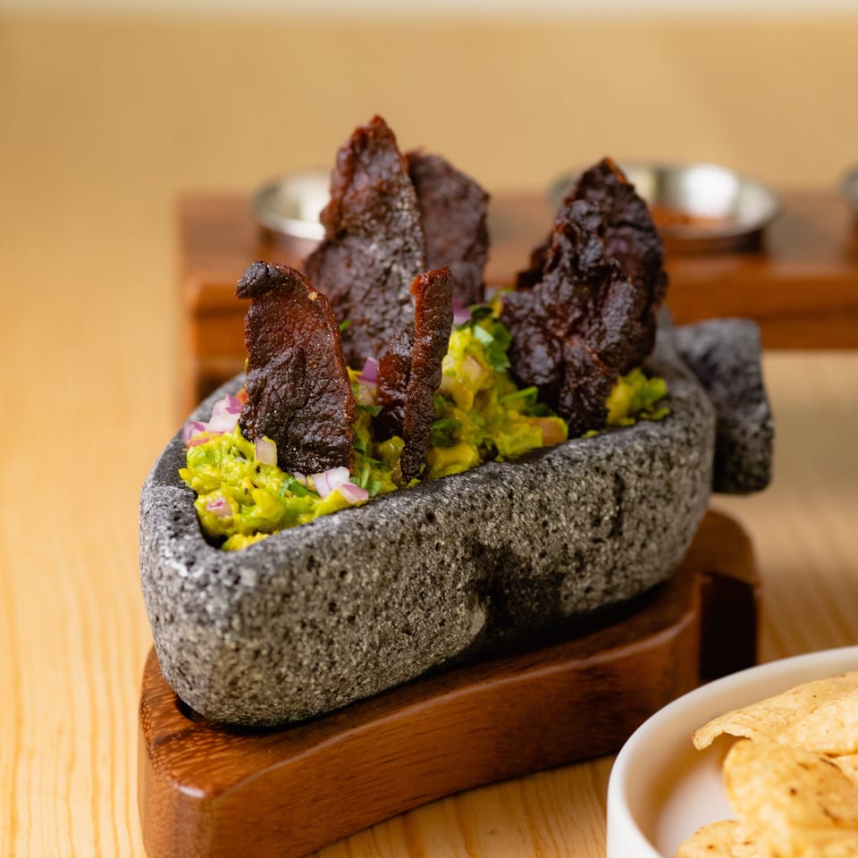 Stone serving dish holds guacamole topped with crispy dark tortilla strips, alongside tortilla chips on a wooden table.