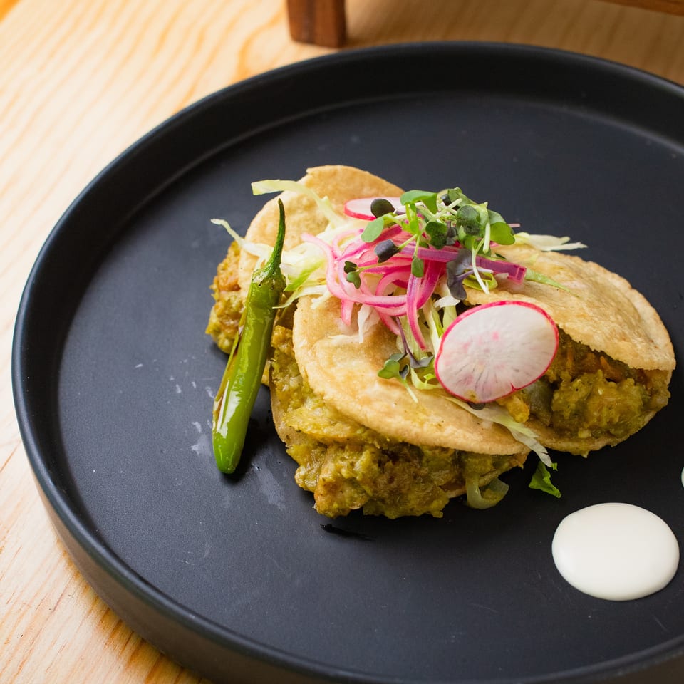 Crispy tacos filled with green sauce-coated filling, topped with pickled onions, radish, microgreens, and served with roasted pepper and cream.