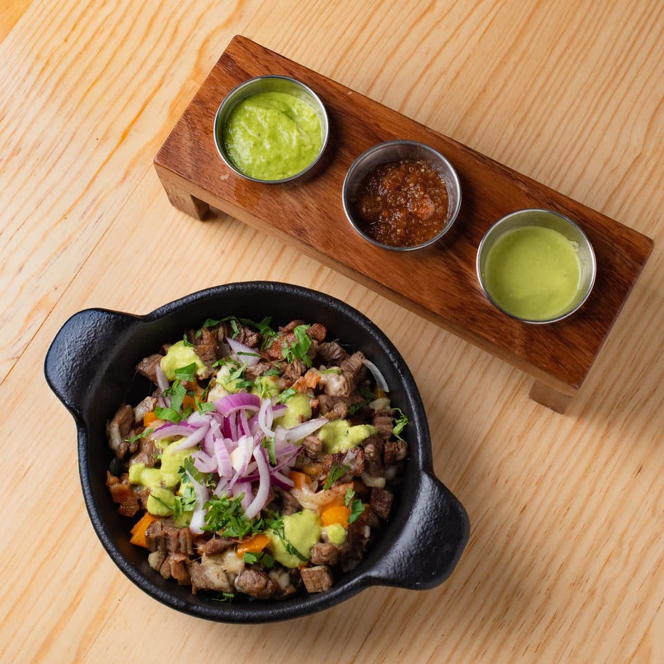 Cast-iron skillet of chopped grilled meat topped with avocado, onions, and cilantro, served with three dipping sauces on a wooden board.
