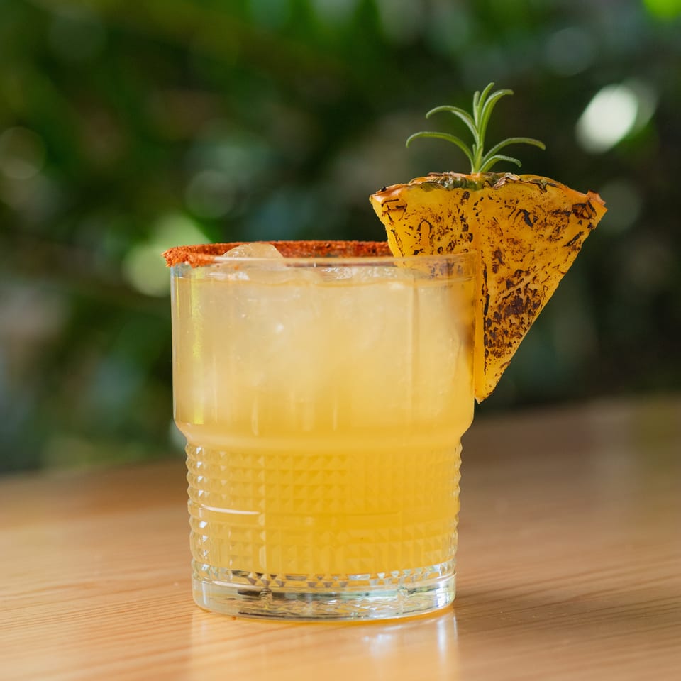 Golden cocktail in a textured glass with a chili-salt rim, pineapple wedge, and rosemary sprig on a wooden table.