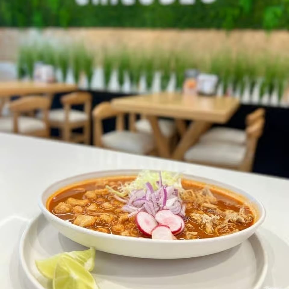 Bowl of reddish Mexican soup topped with cabbage, red onion, radishes, and crispy toppings, served with lime wedges at Chiludos.