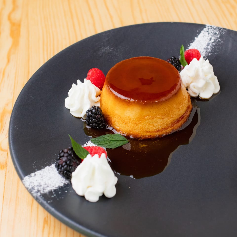 Caramel flan garnished with whipped cream, raspberries, blackberries, mint, and powdered sugar on a black plate.