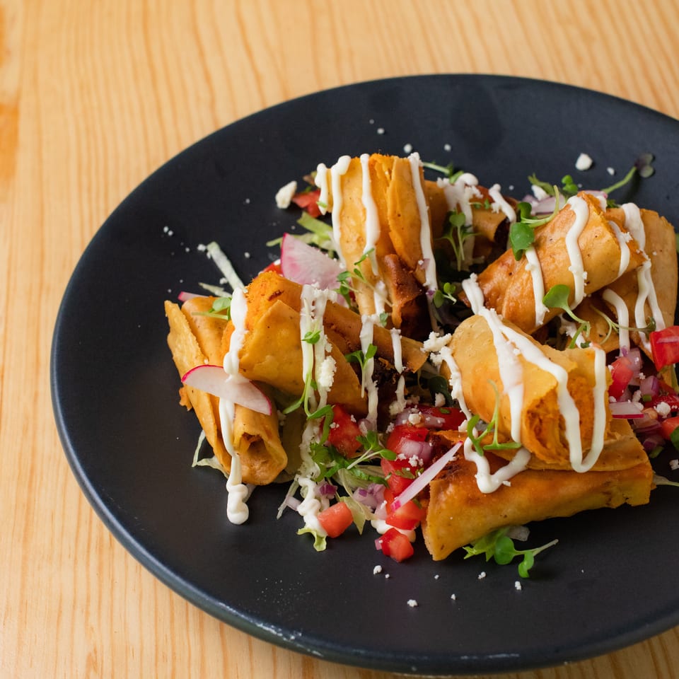 Crispy golden taquitos topped with creamy sauce, fresh vegetables, herbs, and crumbled cheese on a black plate.
