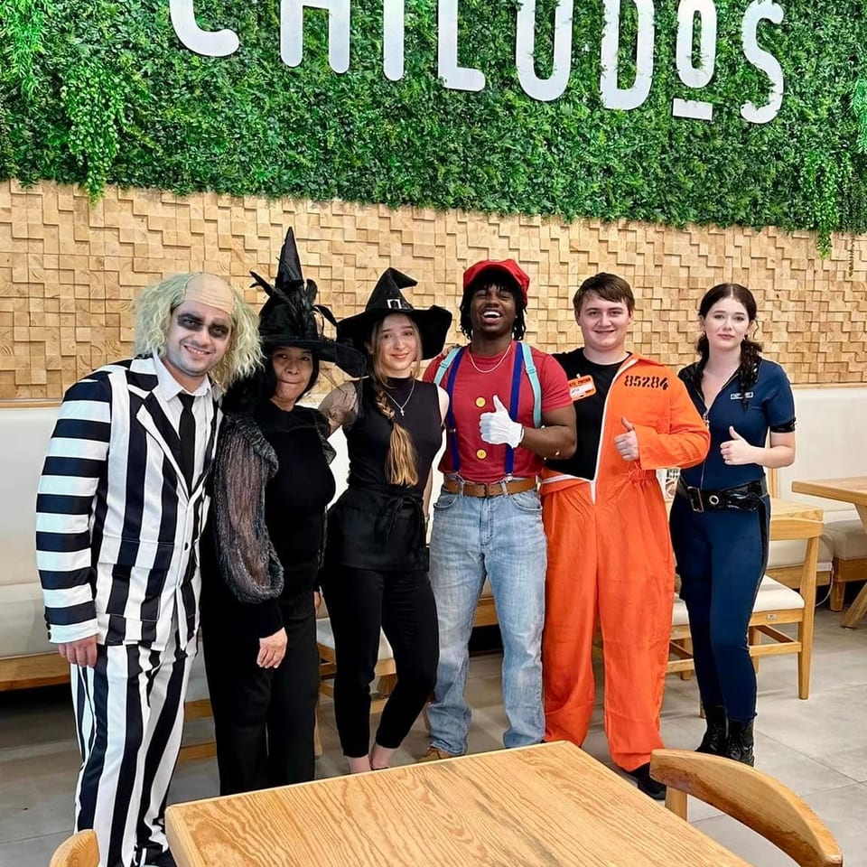 Six people in Halloween costumes pose beneath the CHILUDOS restaurant sign, with tables and chairs in the dining area.