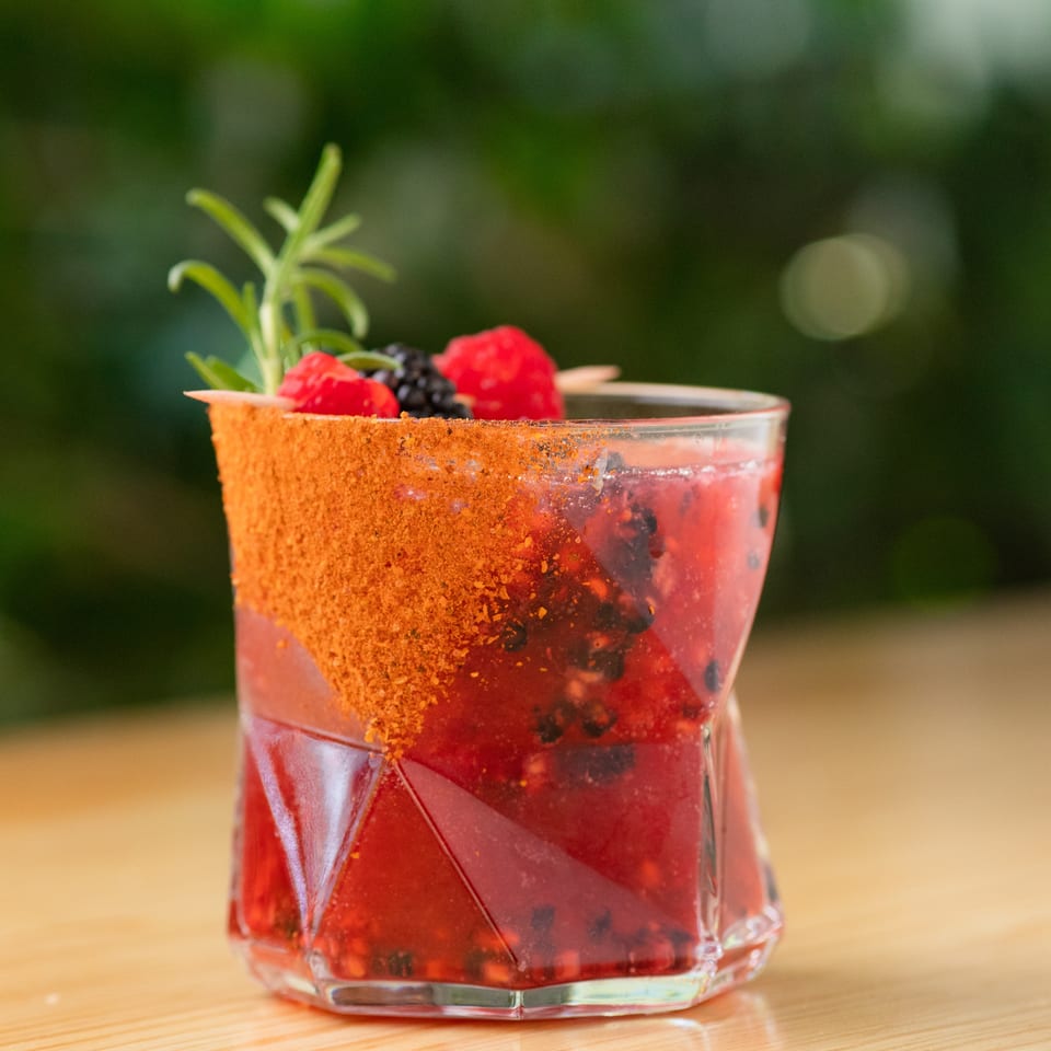 Red berry cocktail with blackberries, raspberries, herbs, and a chili-seasoned rim in a faceted glass on a wooden table.