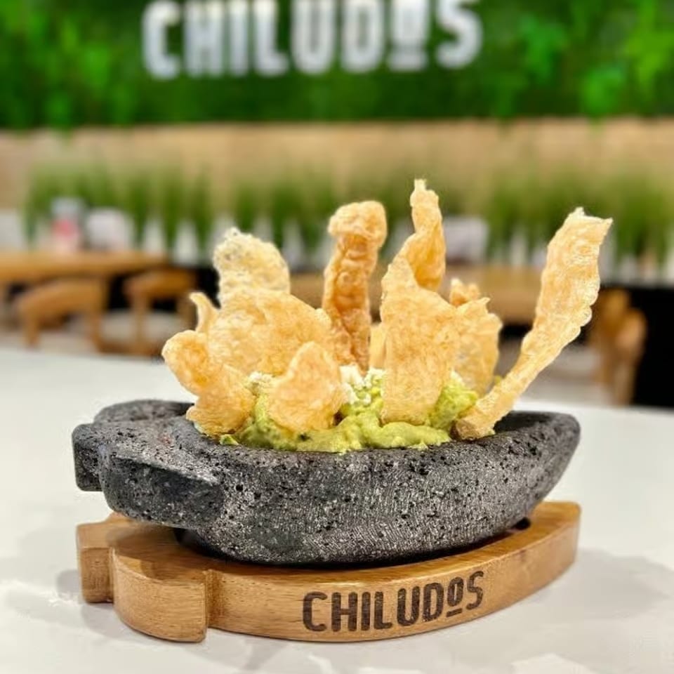 Crispy fried pieces with guacamole served in a black stone bowl on a wooden Chiludos-branded stand.