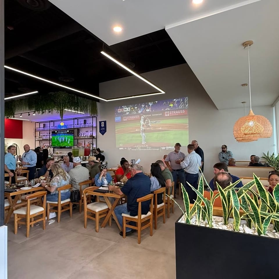 Busy modern restaurant dining room with patrons, bar seating, indoor plants, and a baseball game projected on the wall.