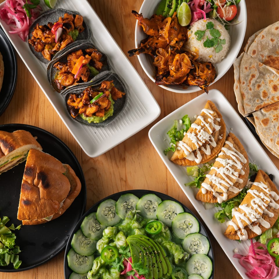 Overhead spread of tacos, quesadillas, grilled shrimp, sandwiches, avocado salad, rice, and colorful pickled vegetable garnishes.